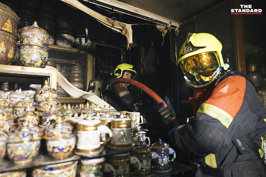 ภาพมุมกว้างเหตุเพลิงไหม้ร้านค้าในตลาดนัดจตุจักรยามค่ำคืน ขณะเจ้าหน้าที่ระดมกำลังฉีดน้ำสกัดเพลิง โดยมีควันไฟพวยพุ่งเหนือหลังคา 1