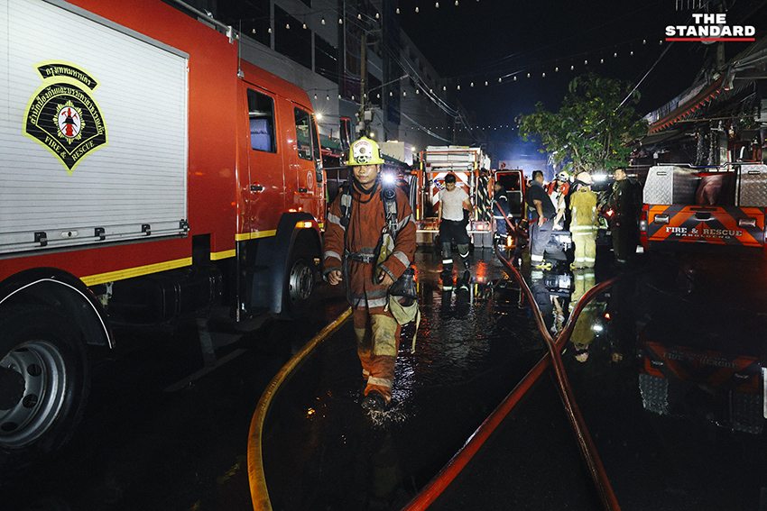 ภาพมุมกว้างเหตุเพลิงไหม้ร้านค้าในตลาดนัดจตุจักรยามค่ำคืน ขณะเจ้าหน้าที่ระดมกำลังฉีดน้ำสกัดเพลิง โดยมีควันไฟพวยพุ่งเหนือหลังคา 10