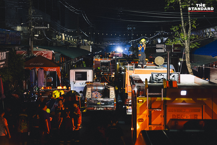 ภาพมุมกว้างเหตุเพลิงไหม้ร้านค้าในตลาดนัดจตุจักรยามค่ำคืน ขณะเจ้าหน้าที่ระดมกำลังฉีดน้ำสกัดเพลิง โดยมีควันไฟพวยพุ่งเหนือหลังคา 15