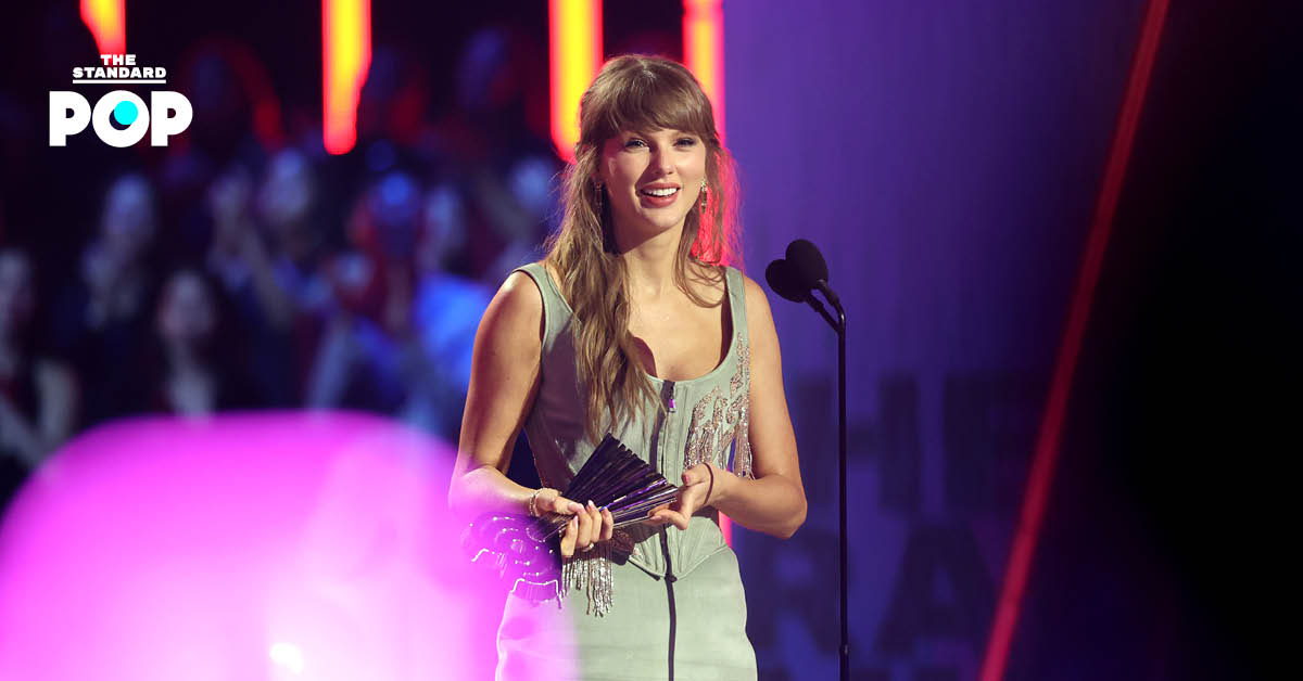 Taylor Swift ในชุดบอดี้สูทสีรุ้งเหลือบมุกสะพายกีตาร์สีชมพูบนเวทีแสงสีม่วง