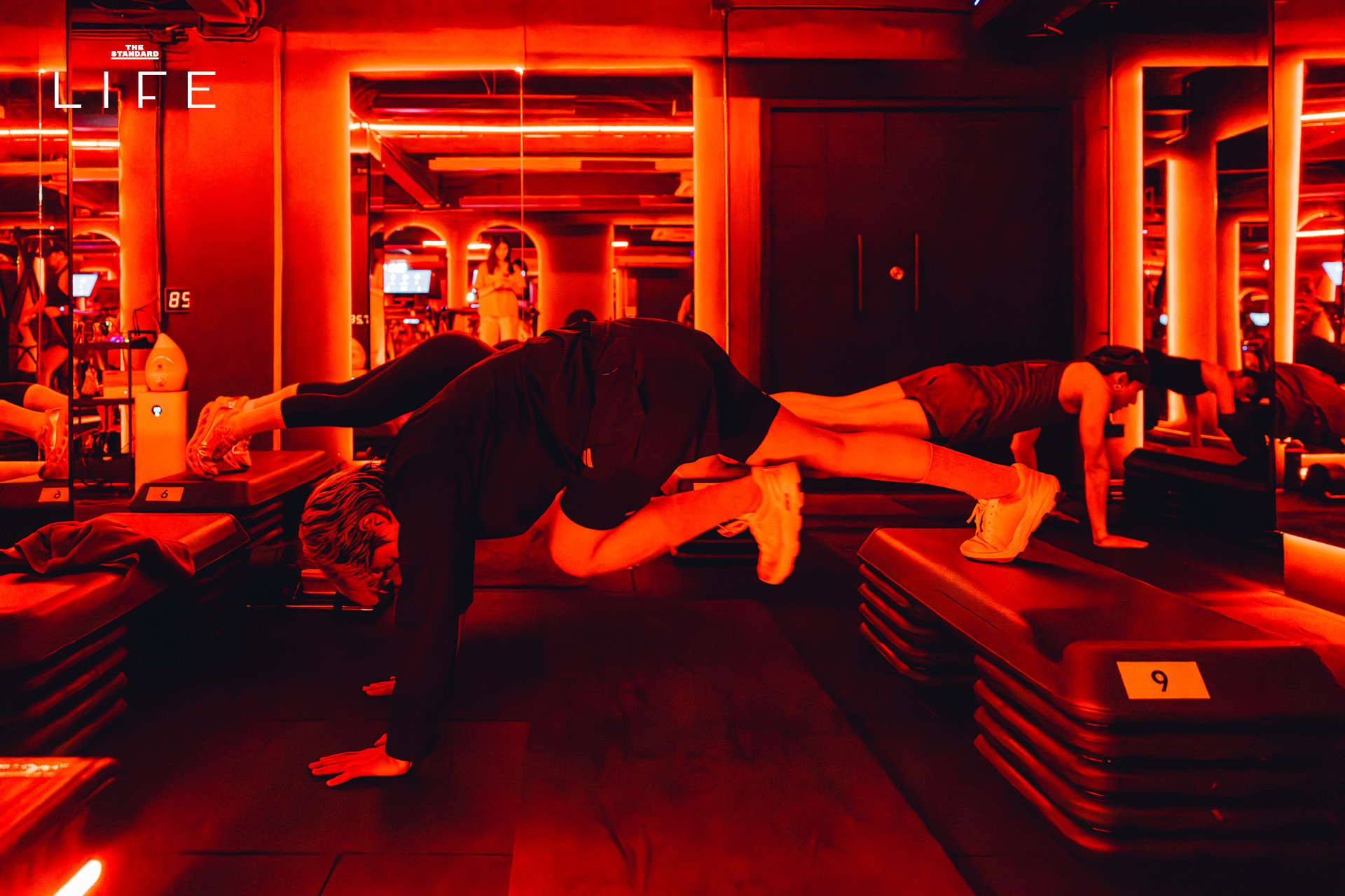 ผู้คนกำลังออกกำลังกายในคลาส HIIT + ASS & ABS ภายในยิม VAULT ที่ตกแต่งด้วยโทนสีแดง 16